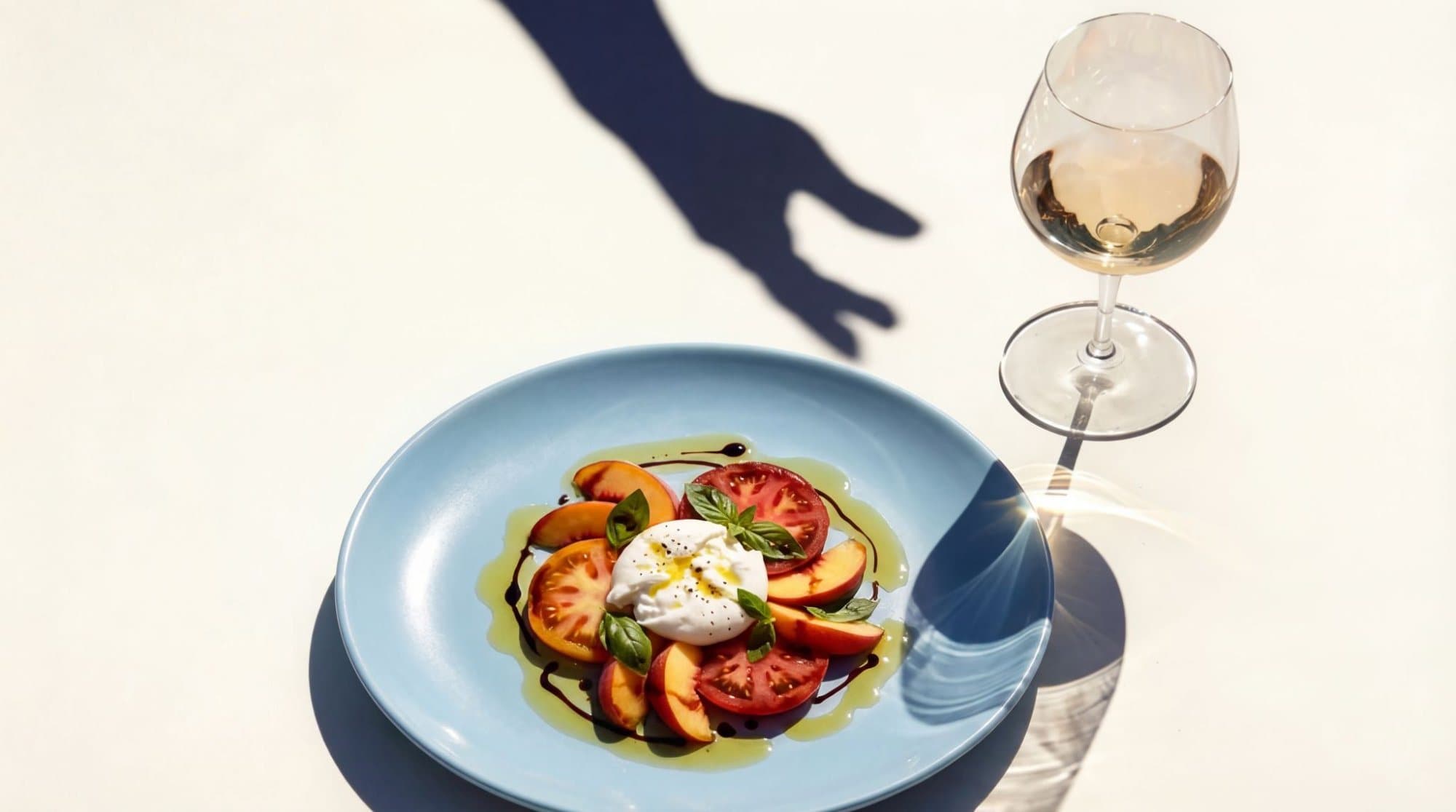 Burrata with peach and tomato on a blue plate beside a glass of white wine in Mediterranean sun